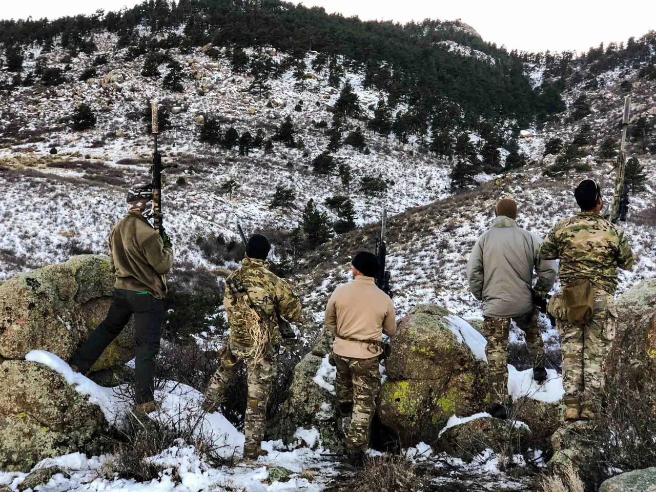 Students with rifles moving through snowy, rocky mountain terrain during a field exercise