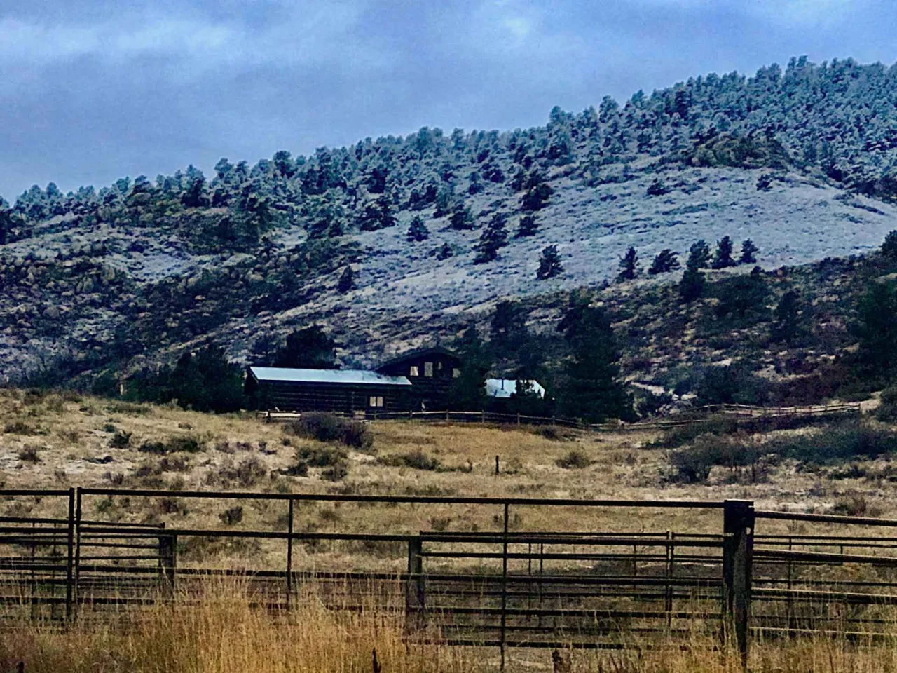 Log ranch buildings and fences below a snow-dusted, pine-covered hillside