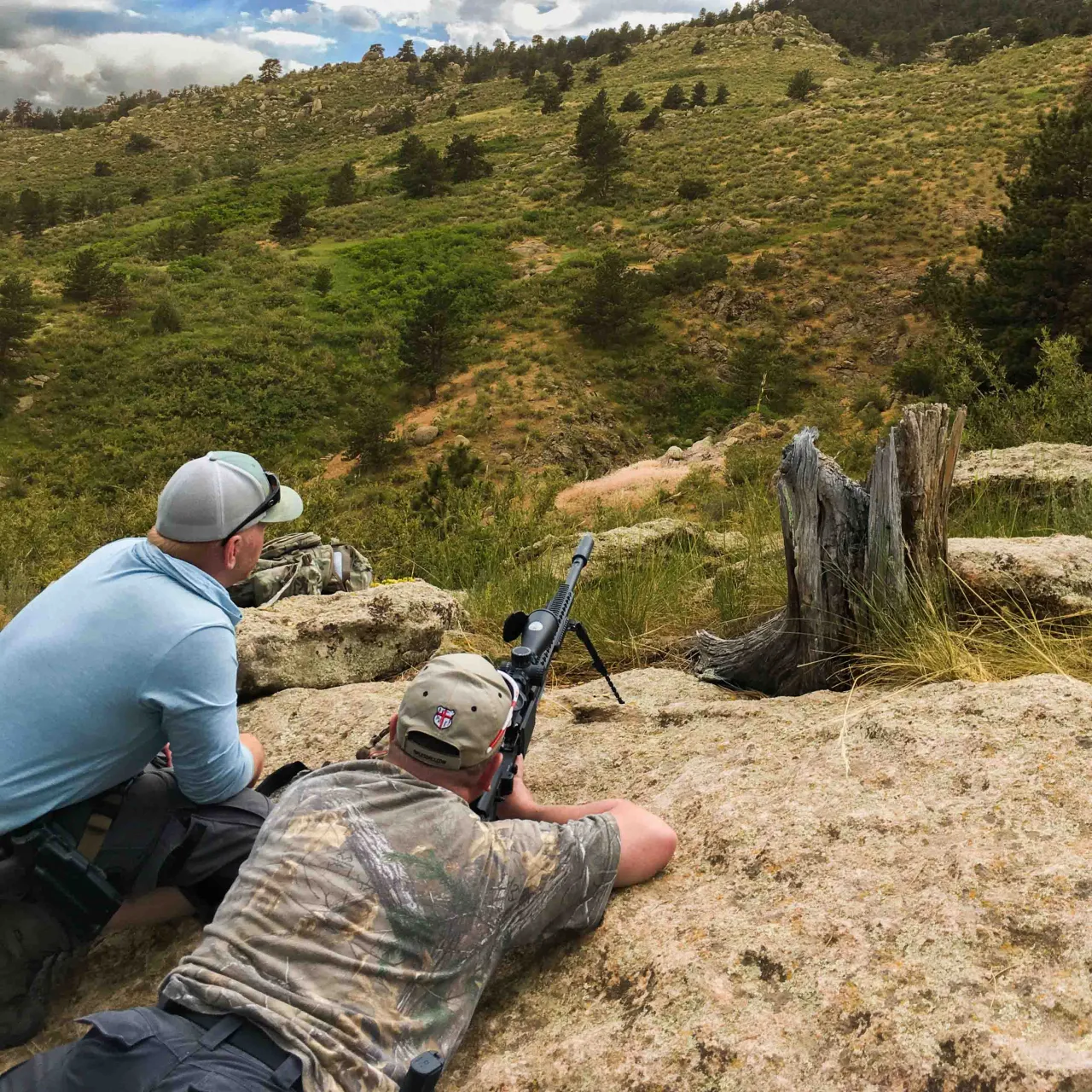 Two students in the prone on a rock outcrop, one aiming a rifle across a green valley
