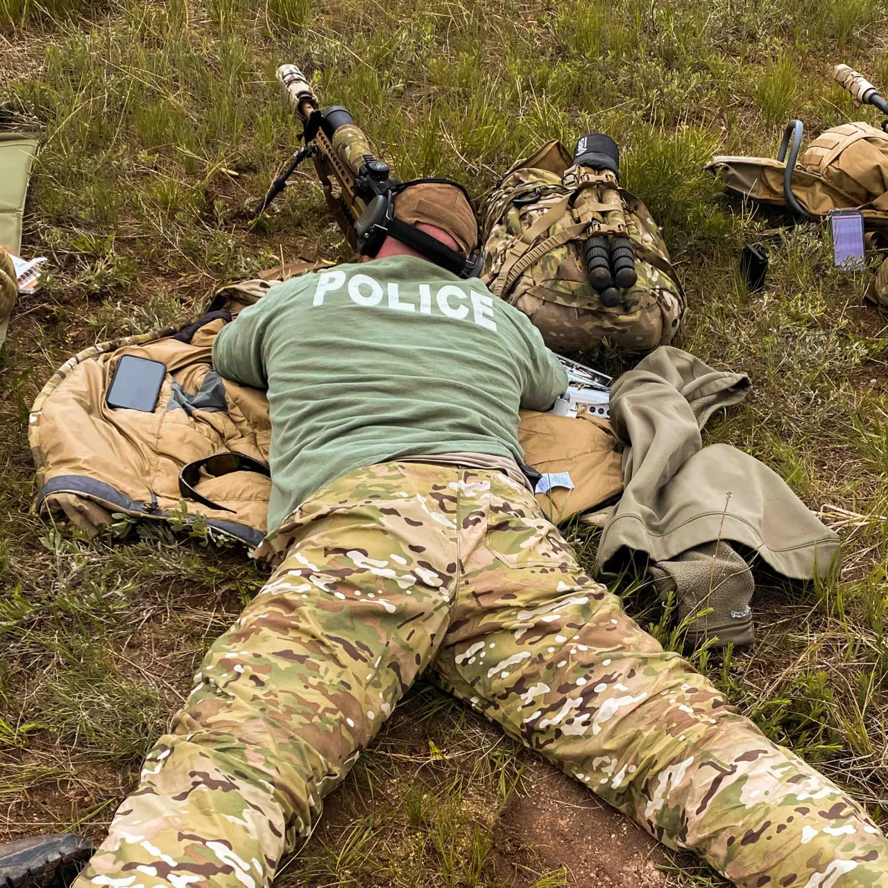 Officer in a Police shirt lying prone behind a rifle on a shooting mat, with gear packs nearby