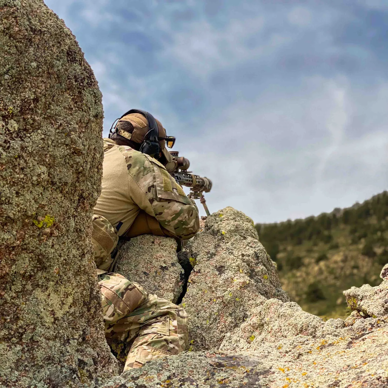 Shooter in camouflage and a helmet aiming a rifle from between tall rocks