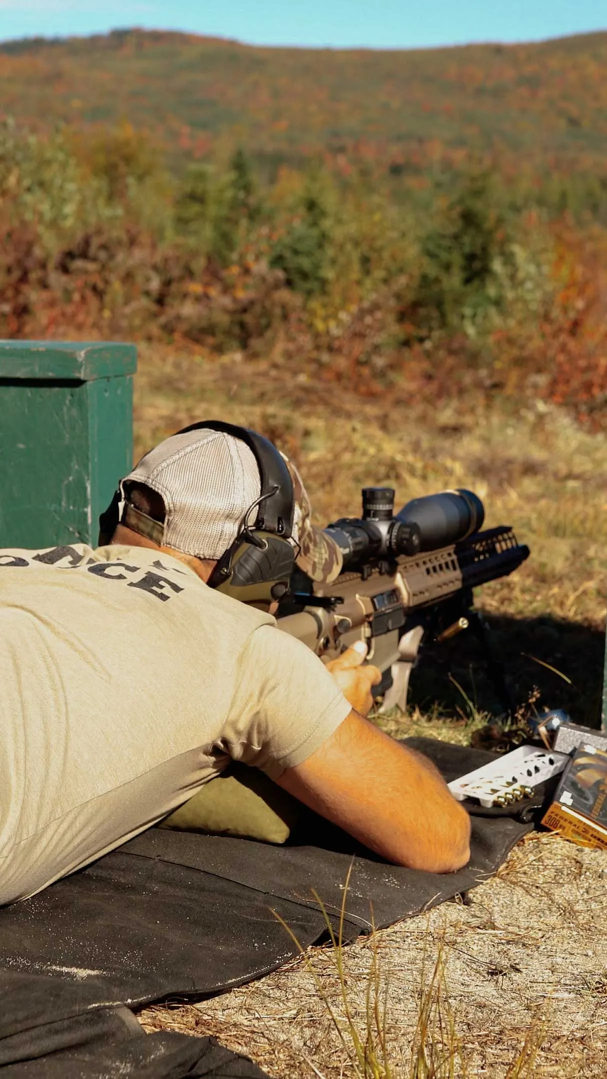 Student lying prone behind a scoped rifle on a shooting mat, with autumn trees behind