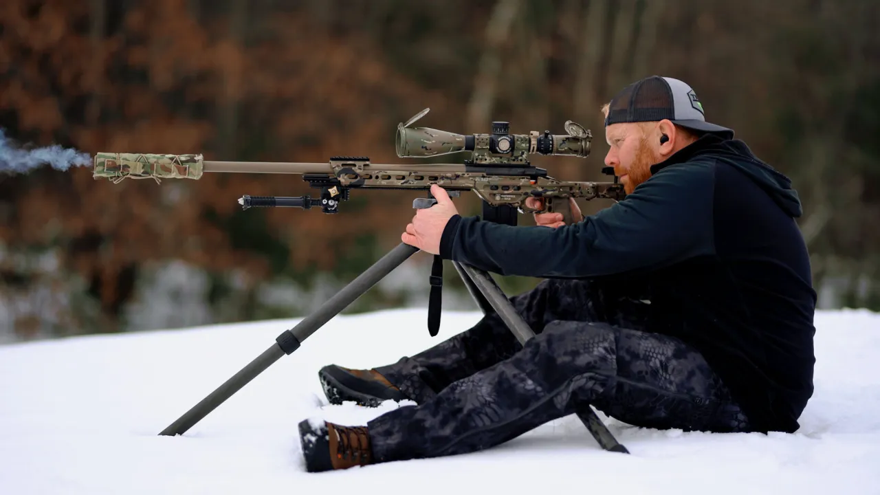 Shooter seated in the snow firing a suppressed rifle supported on a tripod