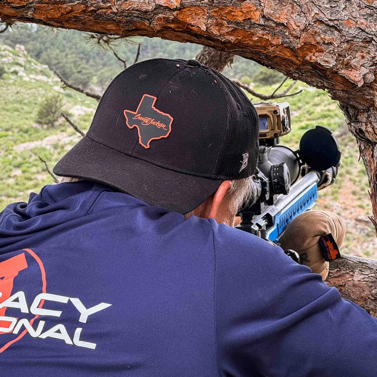 Shooter in a Texas-logo cap aiming a rifle from under a tree branch