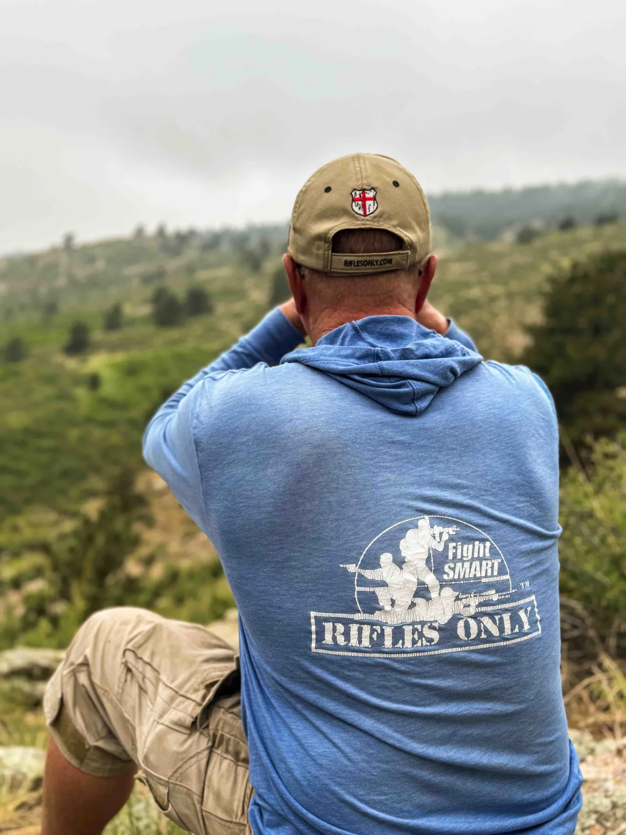 Student in a Rifles Only "Fight Smart" hoodie looking through binoculars over a green valley