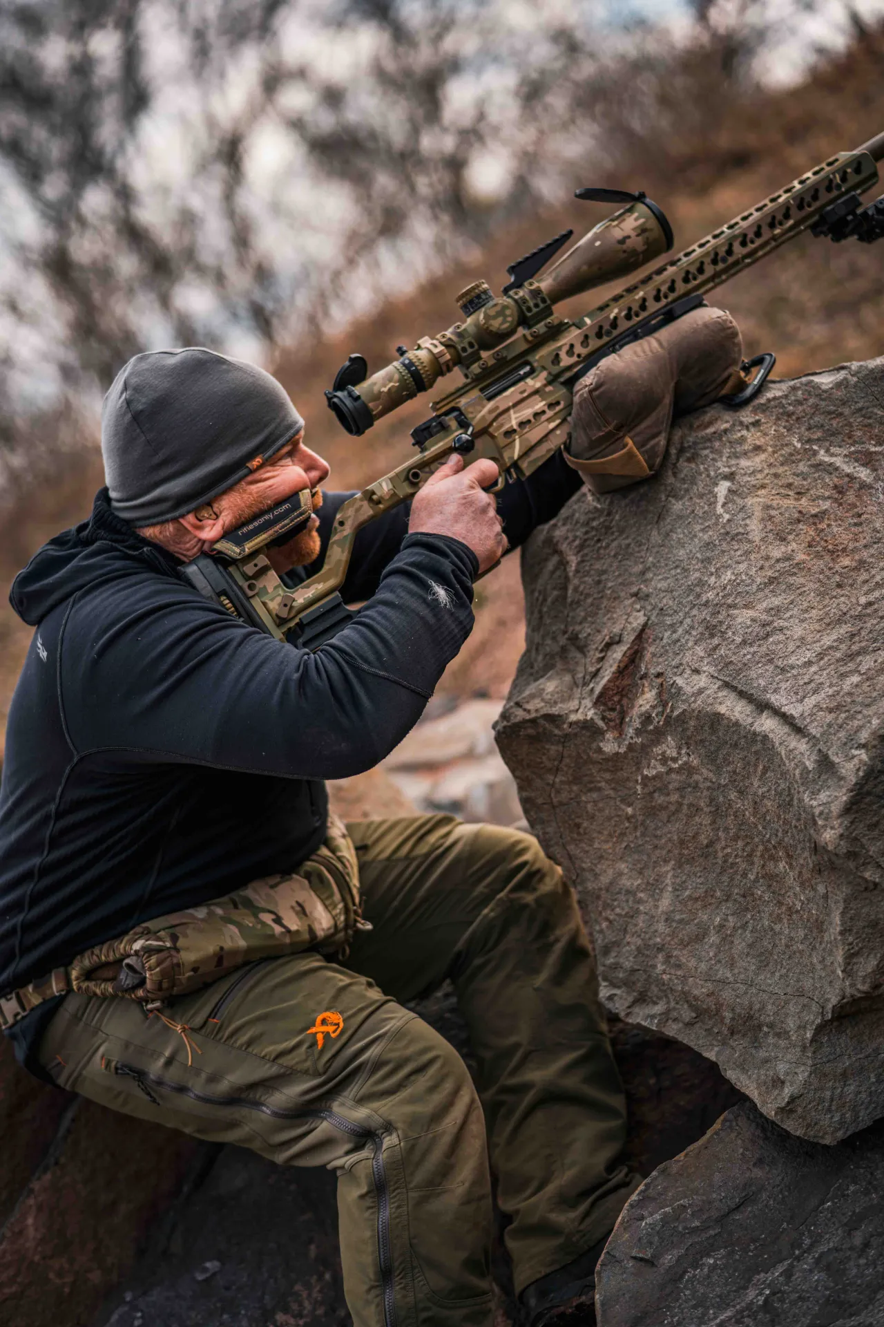 Shooter braced against a rock aiming steeply uphill with a camouflage precision rifle