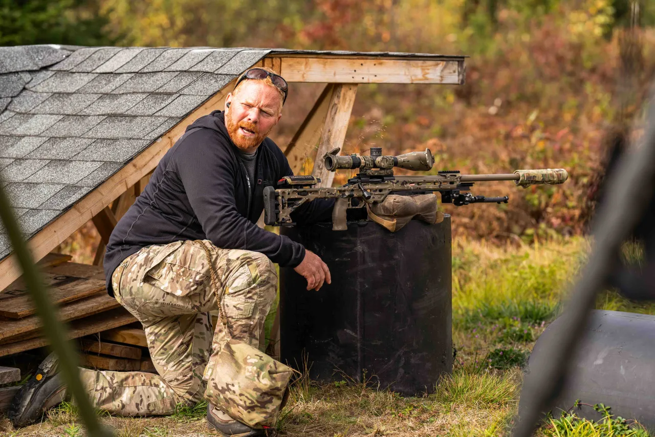 Instructor kneeling beside a precision rifle rested on a barrel, speaking to students