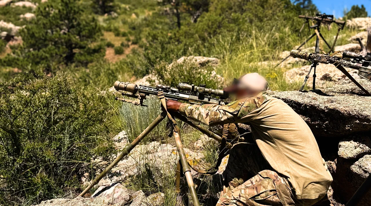 Shooter aiming a rifle on a tripod from rocks on a wooded slope, more rifles set up behind