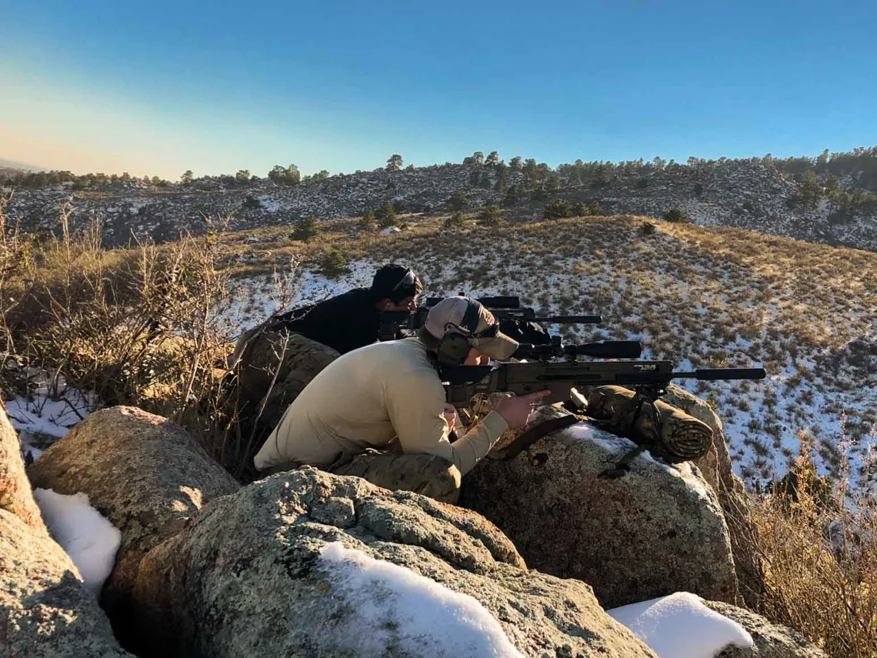 Two students in prone positions behind rocks, aiming precision rifles across a snow-dusted valley
