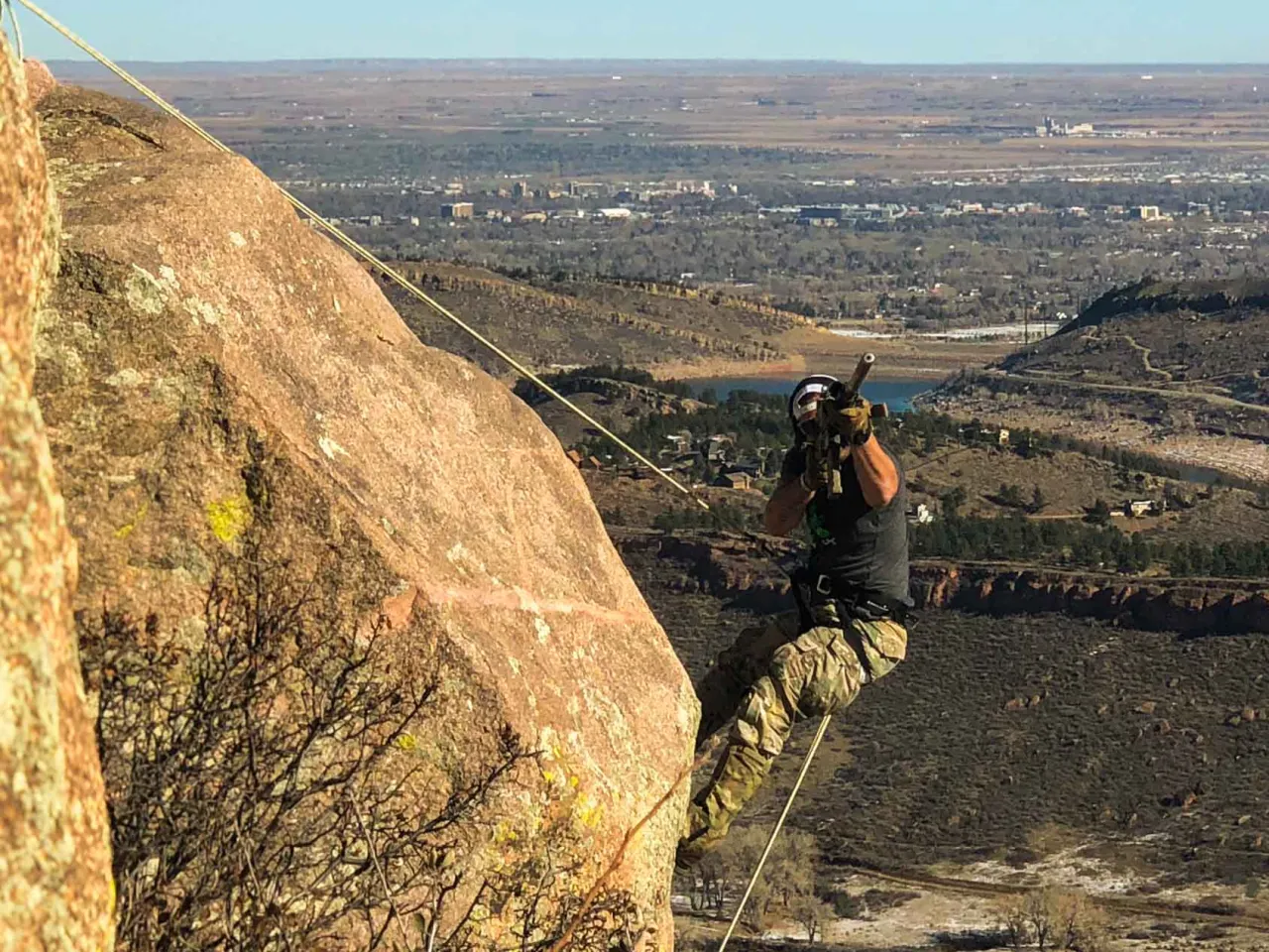 Student rappelling down a large rock face with a rifle slung