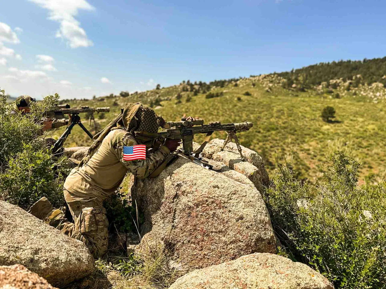 Shooter with an American flag patch aiming a rifle from rocks on a green hillside
