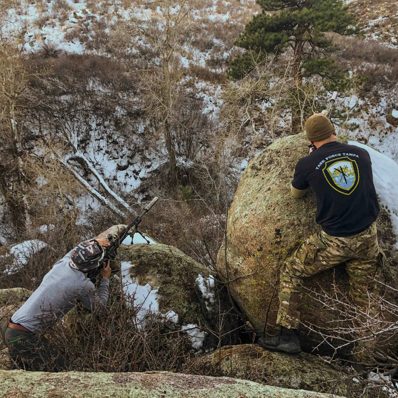 Two shooters taking cover behind a large boulder, one aiming a rifle across the slope
