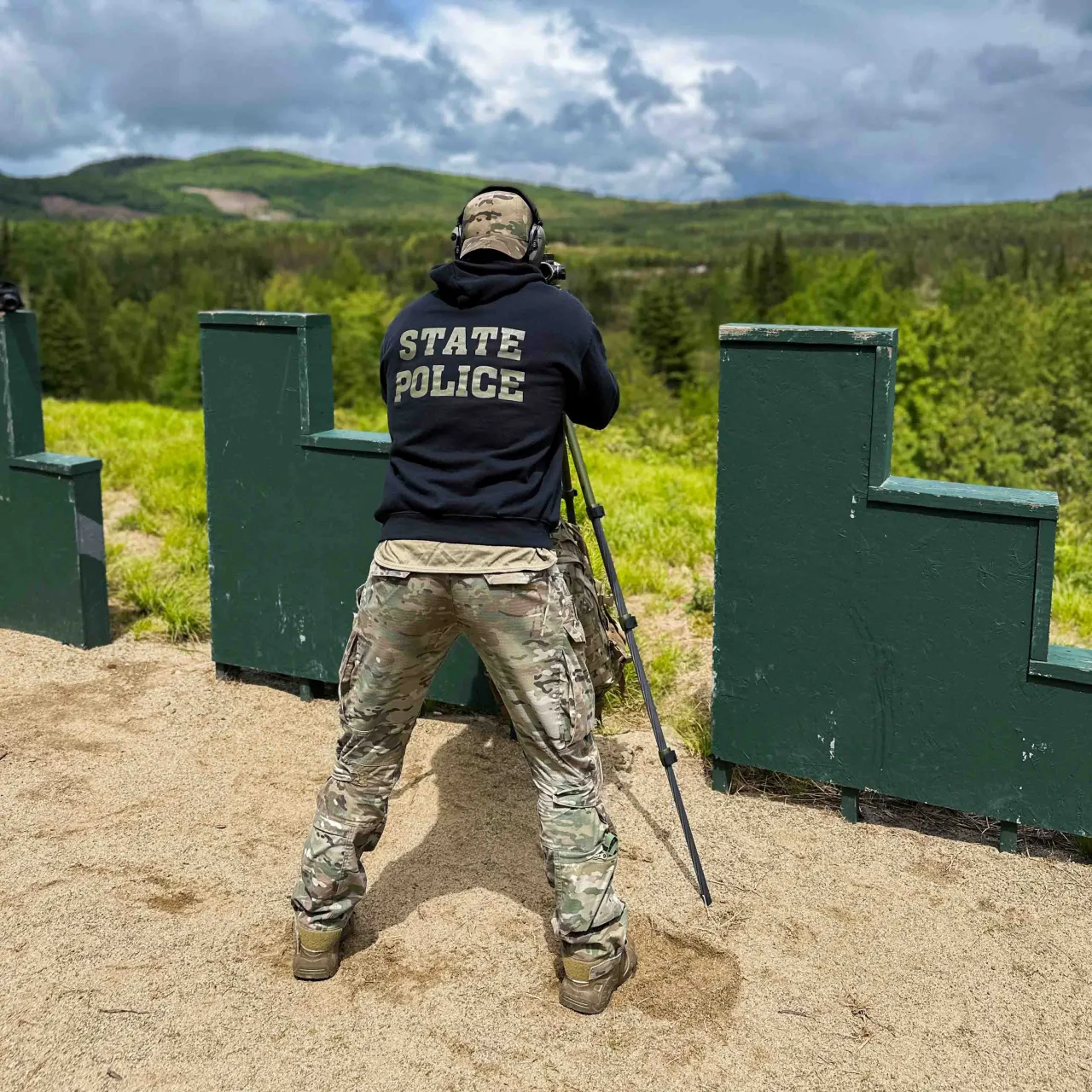Officer in a State Police hoodie aiming from a tripod behind green barricades at the range