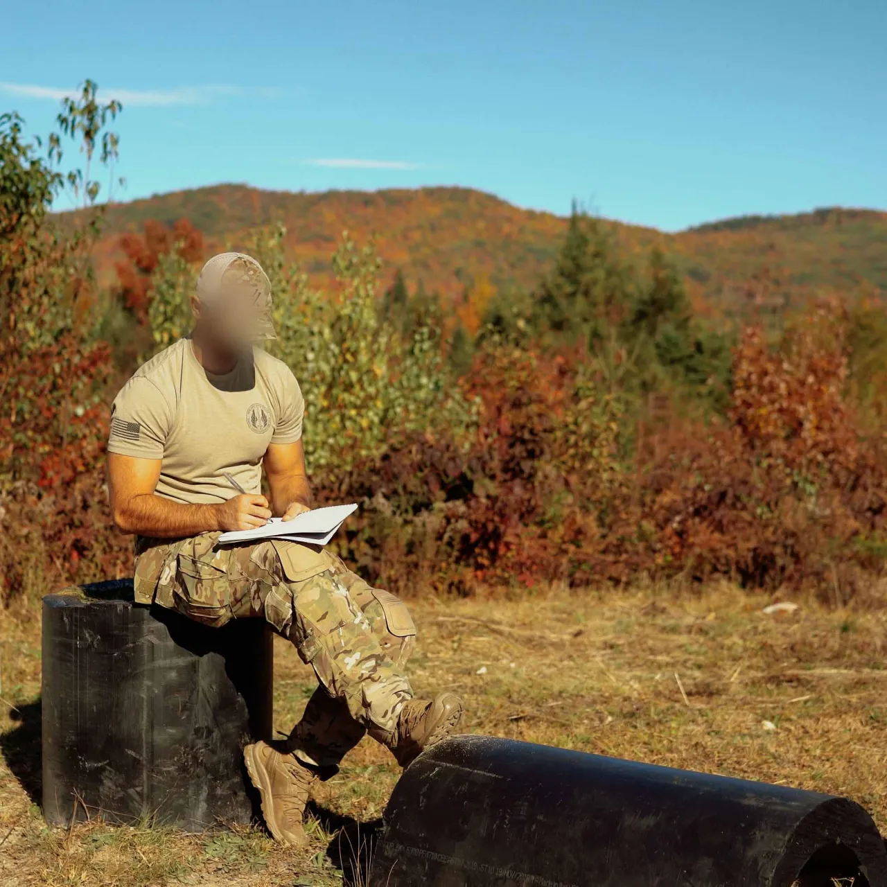 Student sitting on a range barrel writing in a data book, with autumn hills behind