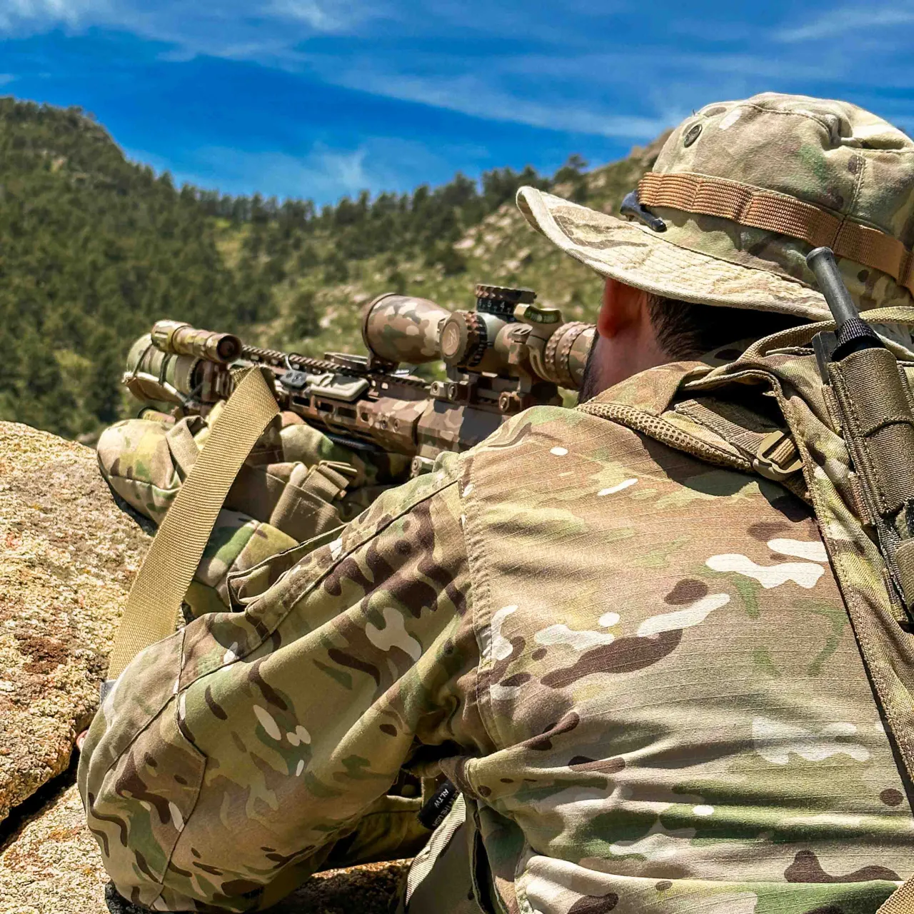Shooter in a boonie hat and camouflage aiming a scoped rifle from a rock on a mountainside