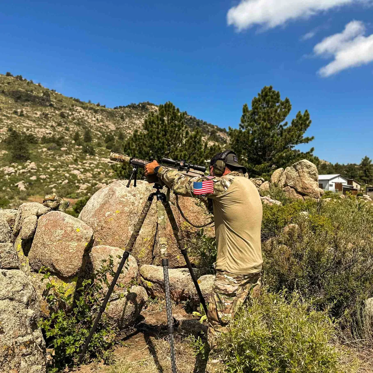 Shooter aiming a rifle on a tripod from a rocky outcrop on a sunny mountainside