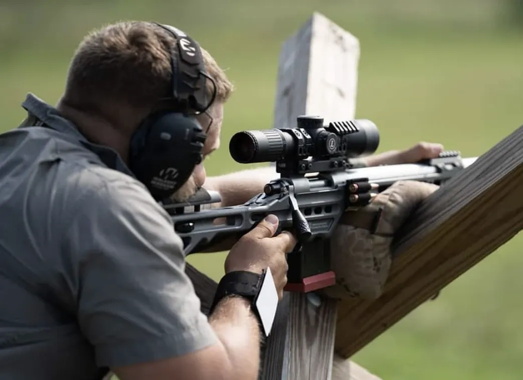 Shooter wearing earmuffs aiming a precision rifle braced on a wooden barricade post