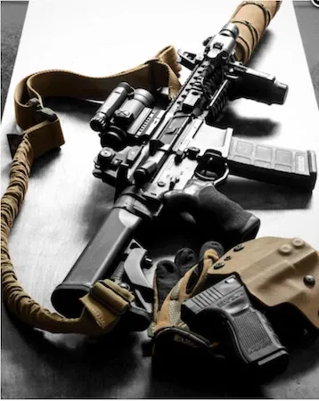 Black-and-white photo of a carbine with an optic, sling, holstered pistol and magazines laid on a table