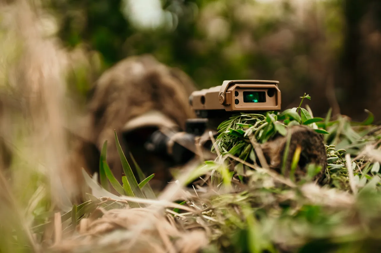 Camouflaged shooter lying in grass behind a rifle fitted with a tan laser rangefinding module