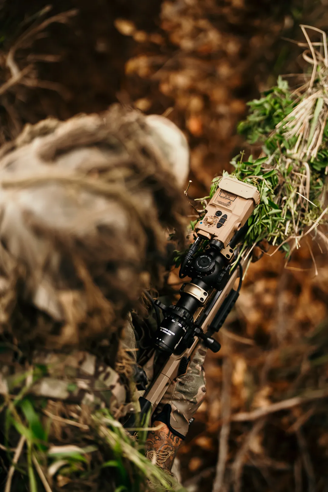 Shooter in a ghillie suit with a rifle-mounted rangefinding module, seen from above in the brush
