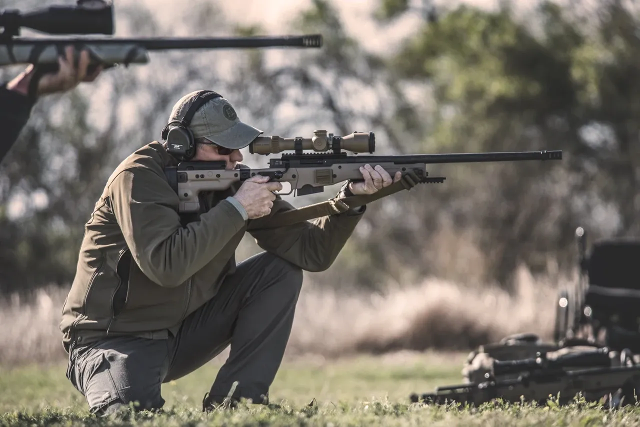 Shooter in a squat position aiming a bolt-action rifle on a grass range