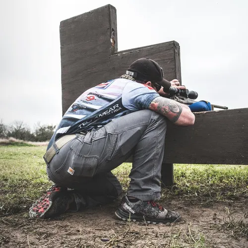 Competitor kneeling behind a tall wooden barricade, aiming a rifle through a notch
