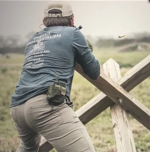 Shooter firing a rifle braced on a wooden X-shaped barricade, spent brass in the air