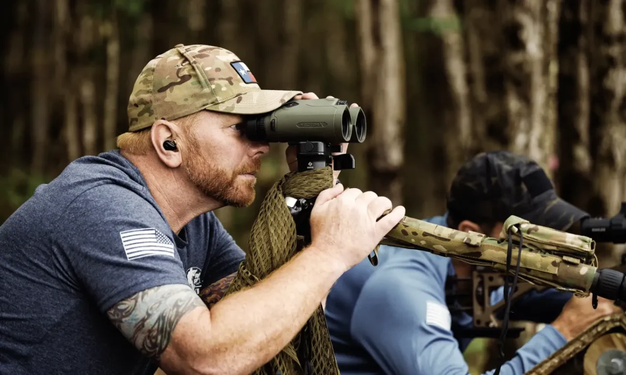Spotter looking through tripod-mounted binoculars beside a shooter behind a rifle