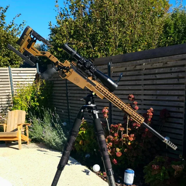 Tan chassis precision rifle with a scope standing on a tripod in a garden