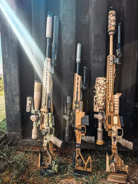 Three tan and camouflage precision rifles leaning against a dark wall in sunlight