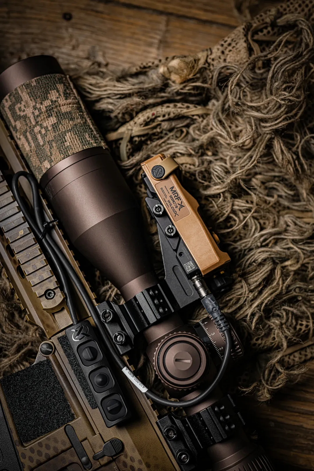 Close-up of a camouflage-wrapped rifle scope with a tan rangefinding module on a ghillie blanket