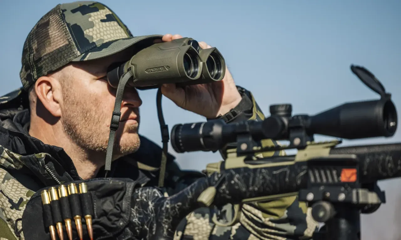 Hunter looking through rangefinding binoculars over a scoped rifle